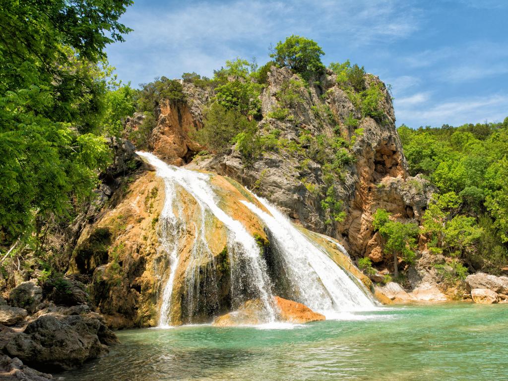 Turner Falls, Oklahoma, USA with water cascading over rocks into a natural pool on a sunny day surrounded by trees.