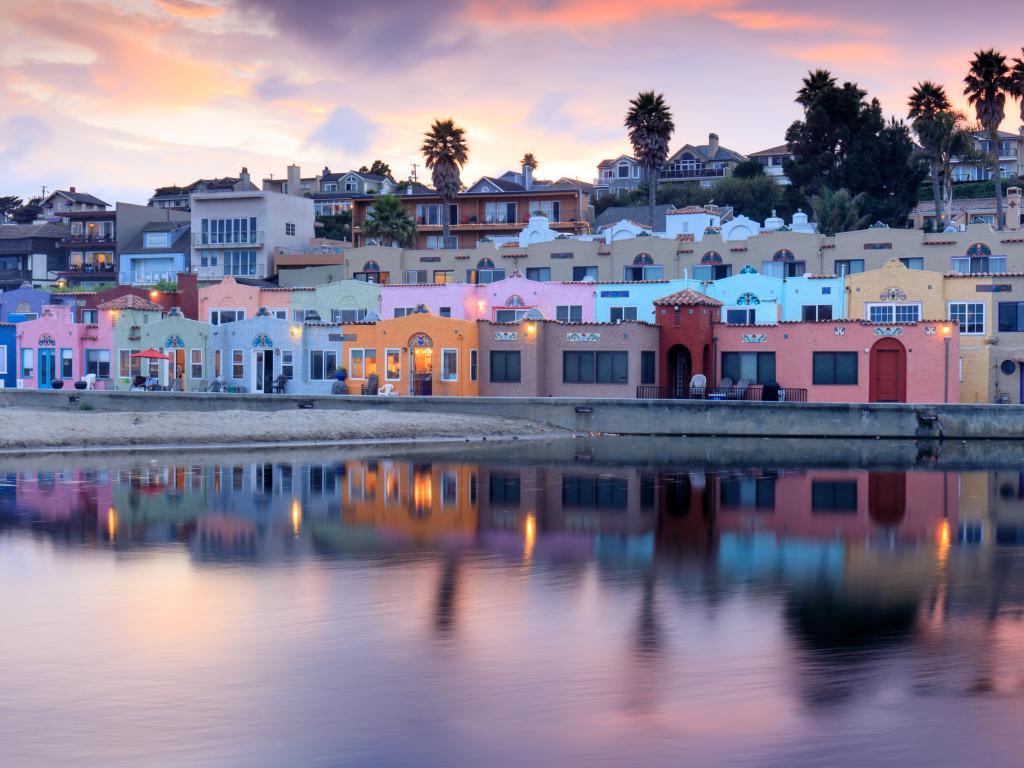 Colourful buildings reflected in still water, with grey and pink sunset sky and palm trees
