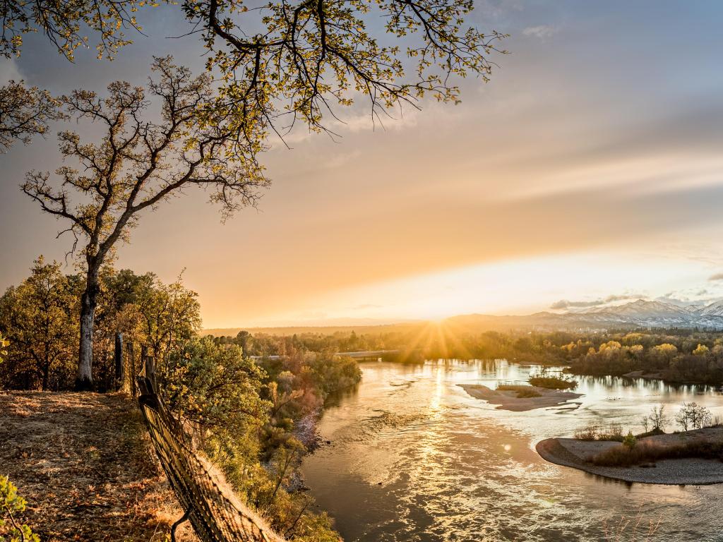 Sacramento River, California taken after a storm with the sun about to set, trees on the edge of the river and mountains faint in the background.