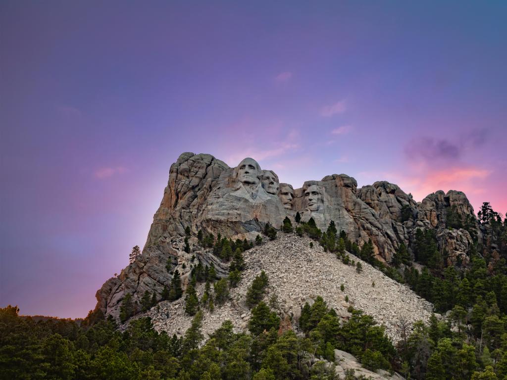 Mount Rushmore, USA with the landscaped Black Hills of Mount Rushmore against a dusk sky of pink and purple and trees in the foreground.