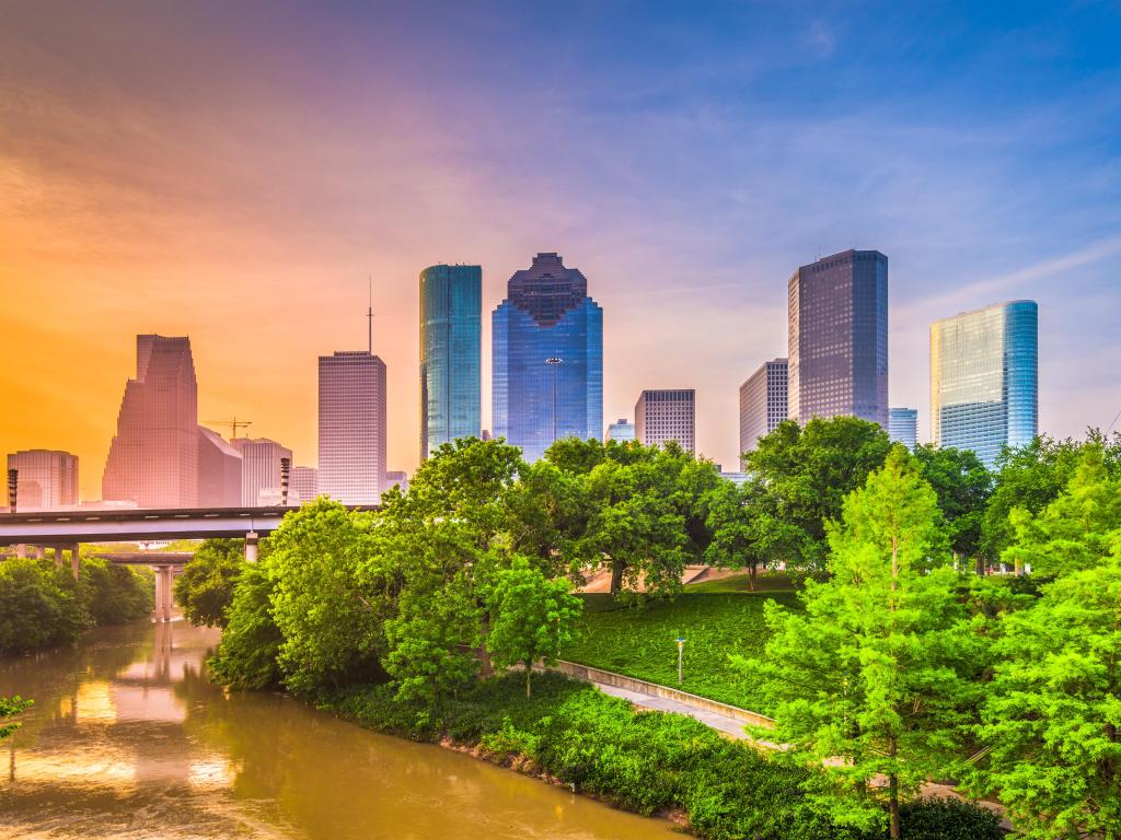 Houston, Texas, USA downtown city skyline against a sunset, with green trees in the foreground and a river leading towards a bridge and the city. 