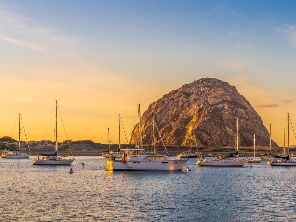 Small white boats in front of beach with sand dunes and large rock rising up out of the ocean, at sunset