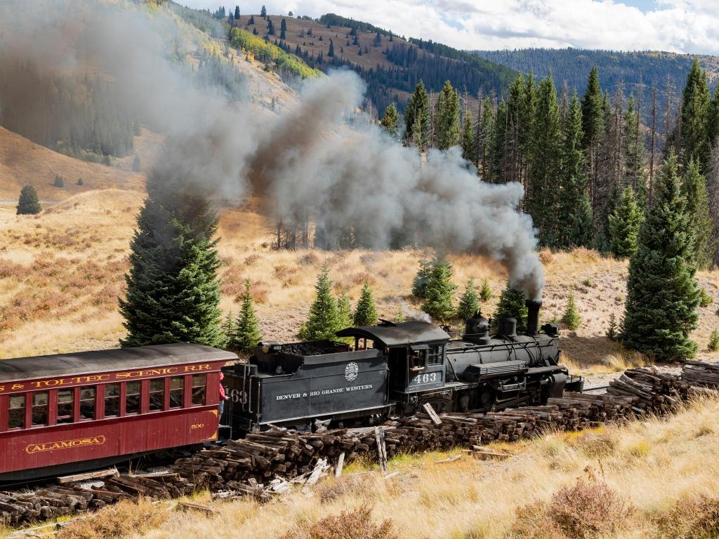 A steam train pulling a dark red carriage across the foothills in New Mexico near Chama