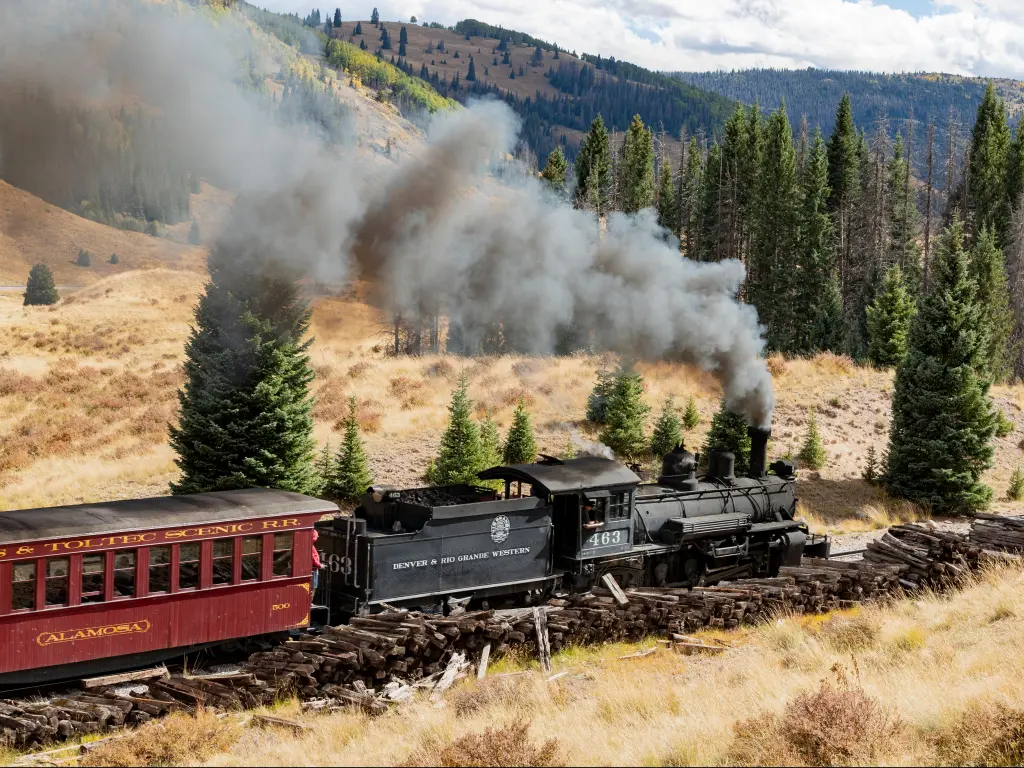 A steam train pulling a dark red carriage across the foothills in New Mexico near Chama