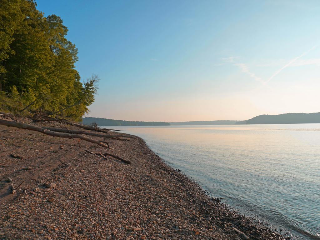 Lake Monroe, IN, USA taken at Rains Burg Beach just before sunset with a clear sky, pebbles in the foreground and calm water, trees in the distance.