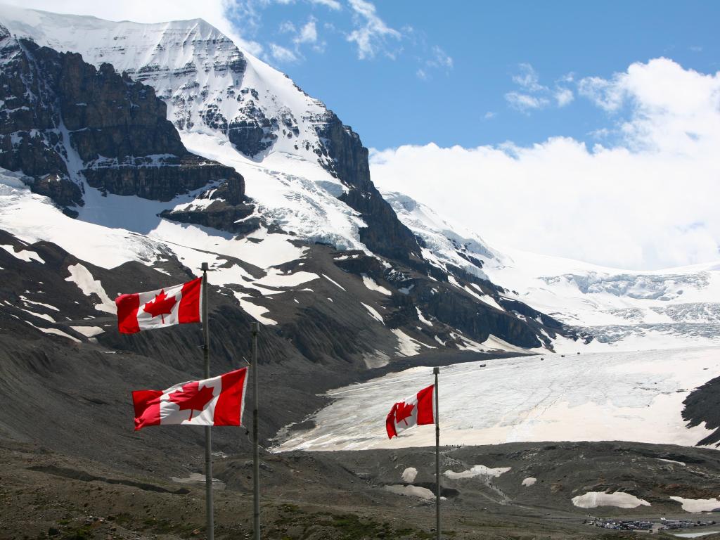 Rugged grey, snow-covered mountain rises up out of the wide icefield, with Maple Leaf flags in the foreground
