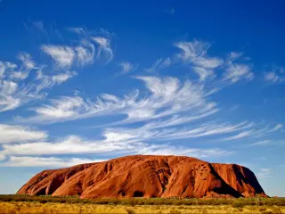 Uluru, Australia showing Ayers Rock surrounded by a blue sky and wispy clouds, with a yellow terrain at the base of the rock. 