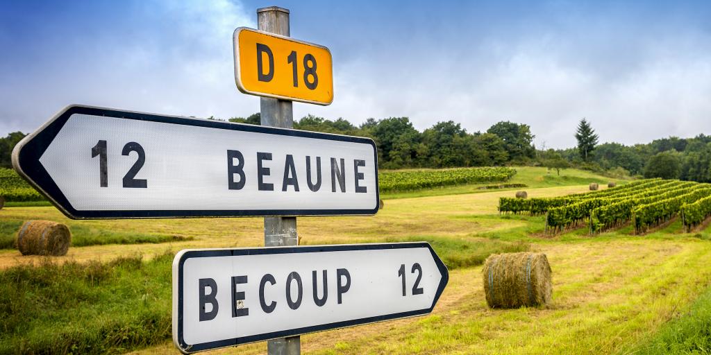 A French road sign pointing to Beaune in one direction and Becoup in another, with hay bales in the background