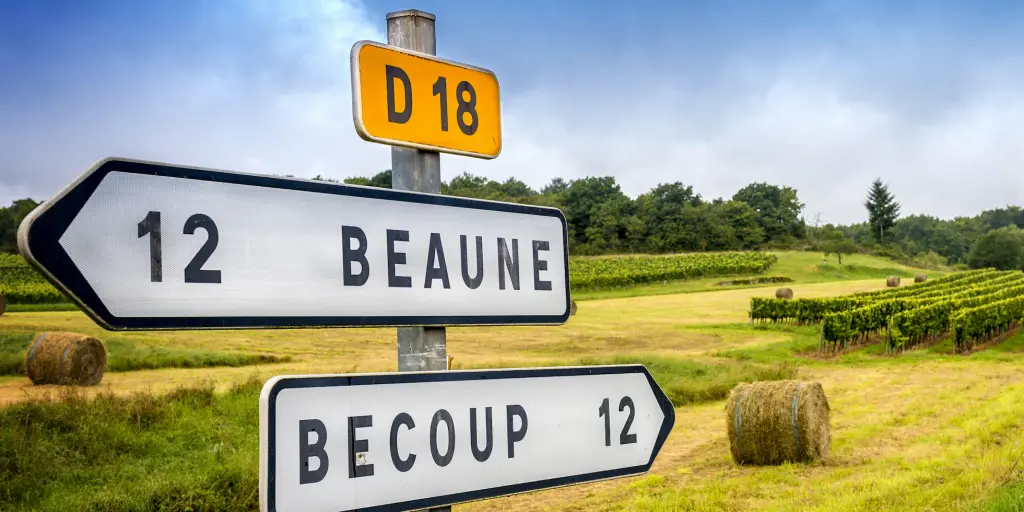 A French road sign pointing to Beaune in one direction and Becoup in another, with hay bales in the background
