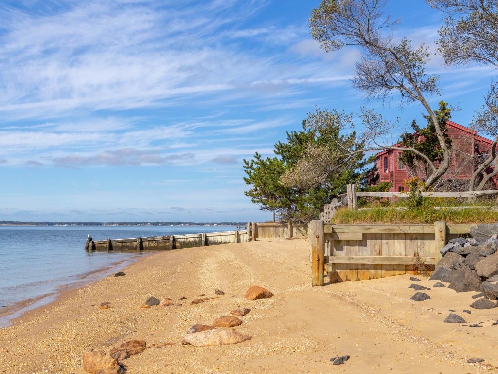 A blue sky with thin sheets of clouds and calm seawater and a tree outside the red house by the beach and red rocks in the sand on a warm sunny day in Shelter Island, New York