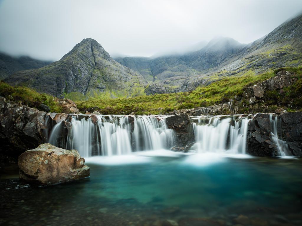 The Fairy Pools, Glen Brittle, Skye, Scotland with a pool in the foreground and waterfall, against a rocky mountain on a foggy day.
