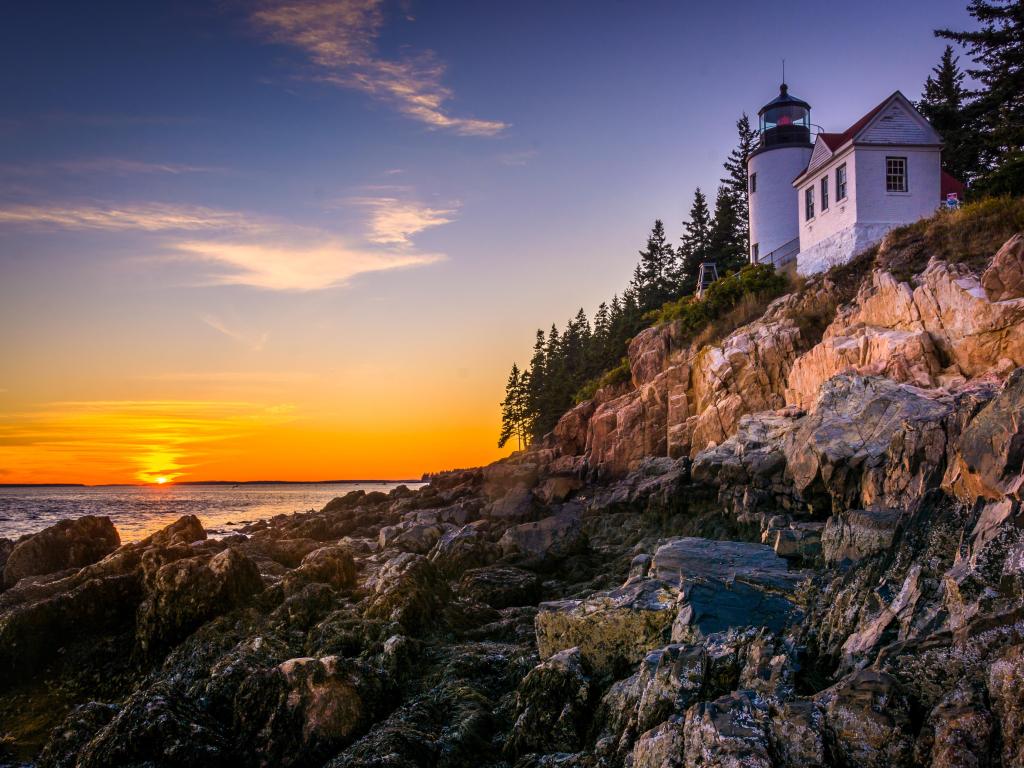 Lighthouse on rocky cliff beside sea with sun setting on the horizon