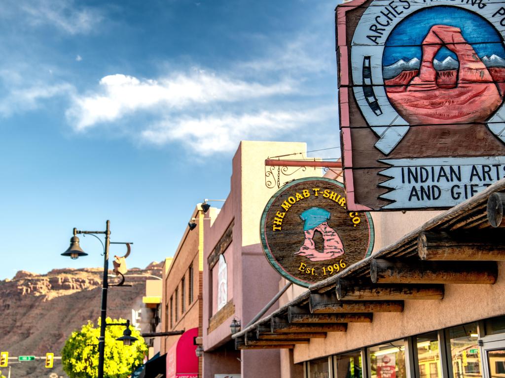 Close up view of colorful store signs in downtown Moab, set against a blue sky with wispy clouds and red rocks in the distance behind