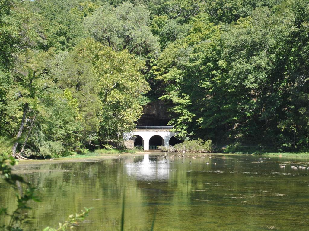 Dunbar Cave Entrance. The photo depicts lush green trees around the entrance area over a lake. 