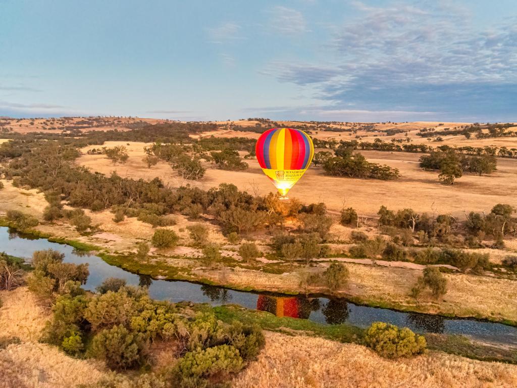 Hot air balloon floating above river across dry landscape with some trees