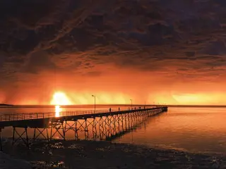  Cloudy overcast sky burnt to orange by sun reflecting in still low tide waters, timber jetty sticking out into the water, beach with trees to one side