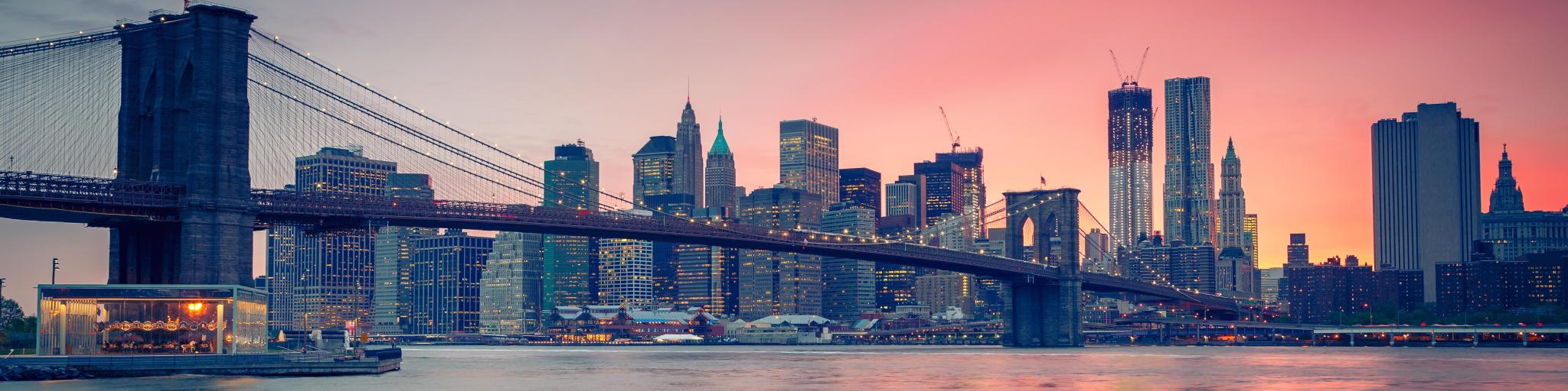 New York City, USA with Brooklyn Bridge at dusk, the city skyline in the distance and a red hue in the sky.