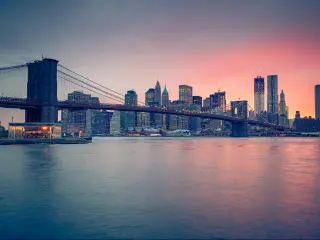 New York City, USA with Brooklyn Bridge at dusk, the city skyline in the distance and a red hue in the sky.
