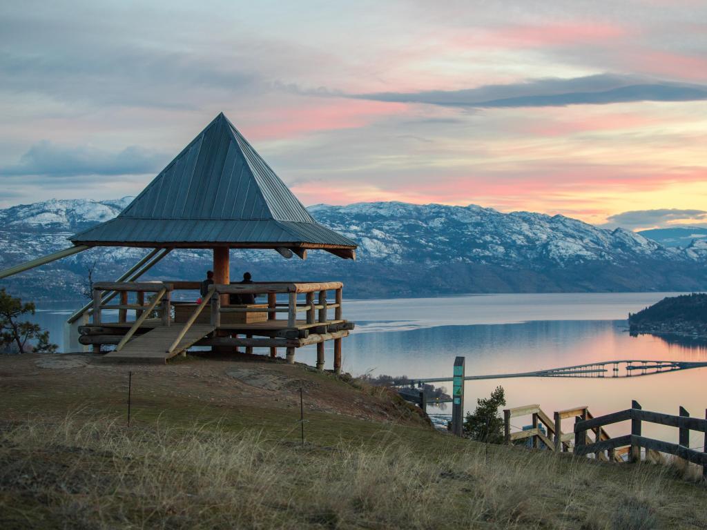 Kelowna, BC, Canada with a landscape photo of a sunset over Okanagan Lake, reflections on the water and snow-capped mountains in the distance. 