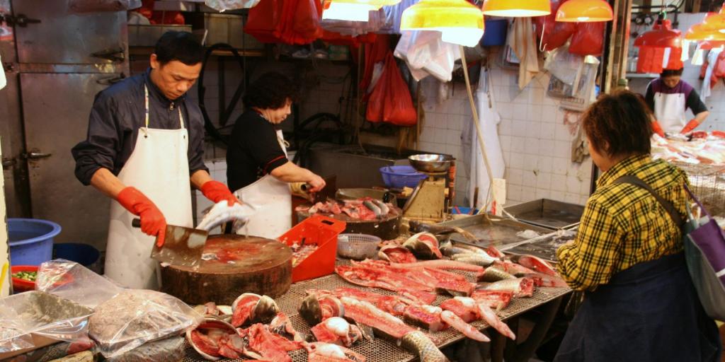 A man gutting a fish at a fish stall in a Hong Kong street market, with a woman cooking fish in the background