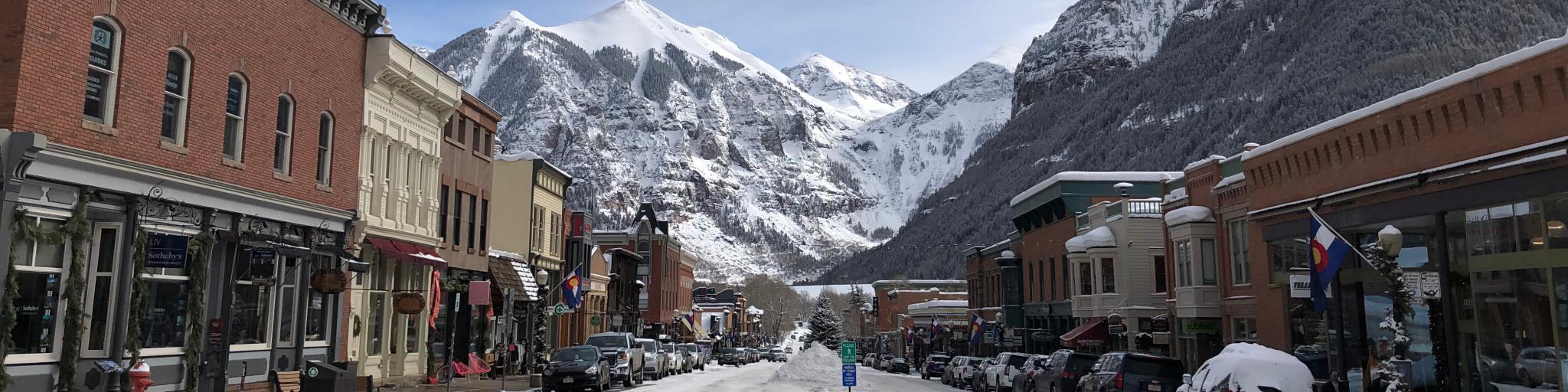 A street view in Telluride, Colorado where snow is on the road and some buildings are on the both sides of the road. The San Juan mountain can be seen on the background.t.