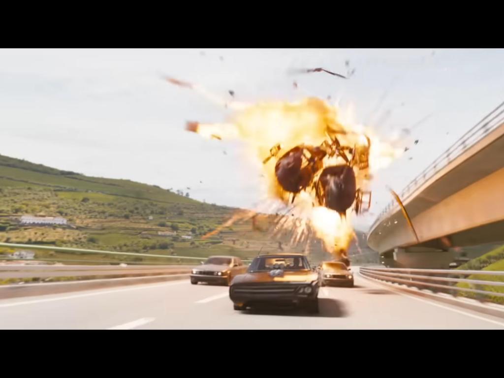 A black car is being chased by other cars on a highway with two helicopters crashing and exploding in the background