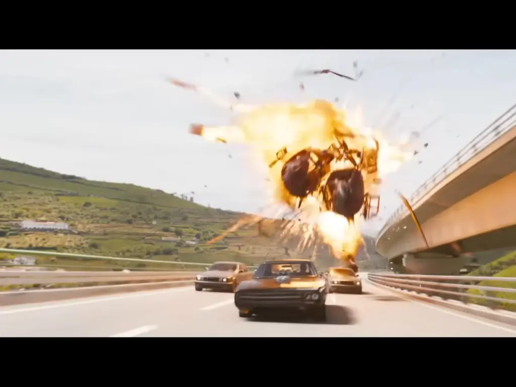A black car is being chased by other cars on a highway with two helicopters crashing and exploding in the background