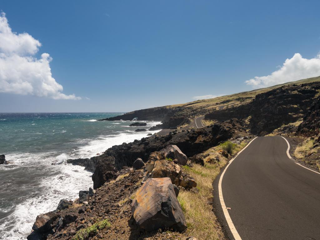 Winding road with a cliff on its left and ocean waves crashing against the rocks on a sunny day
