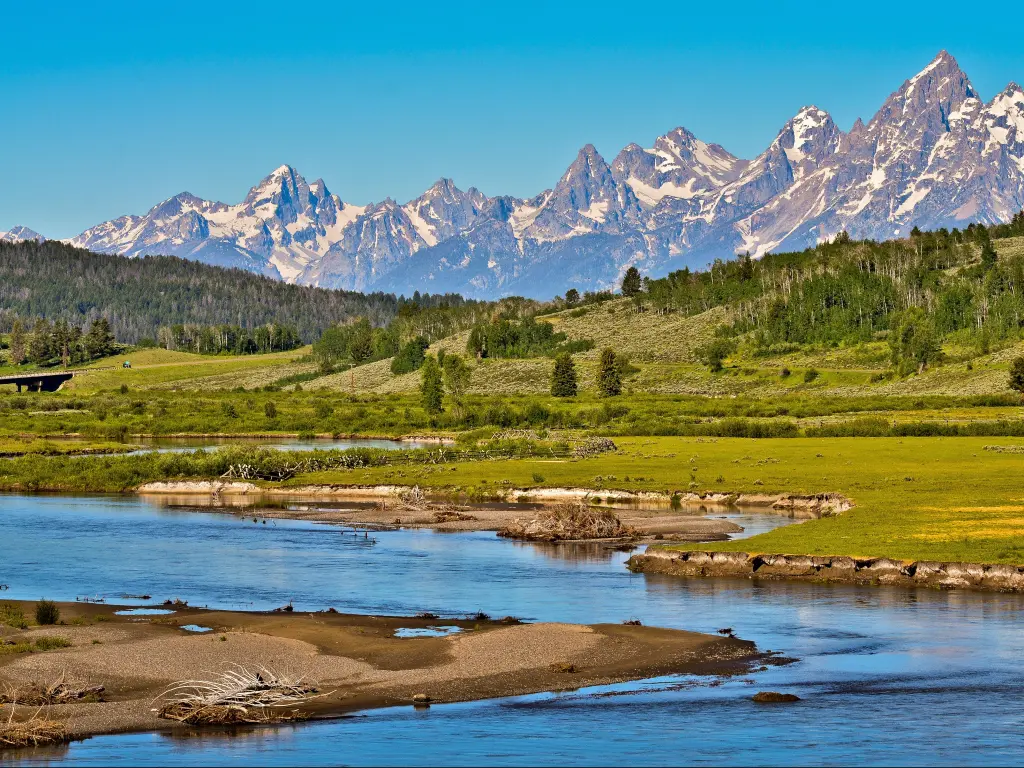 Grand Teton mountain range, partly covered in snow, with green forest, field, and small river in the foreground. It was taken on a sunny day in summer at the gateway to Grand Teton national park.