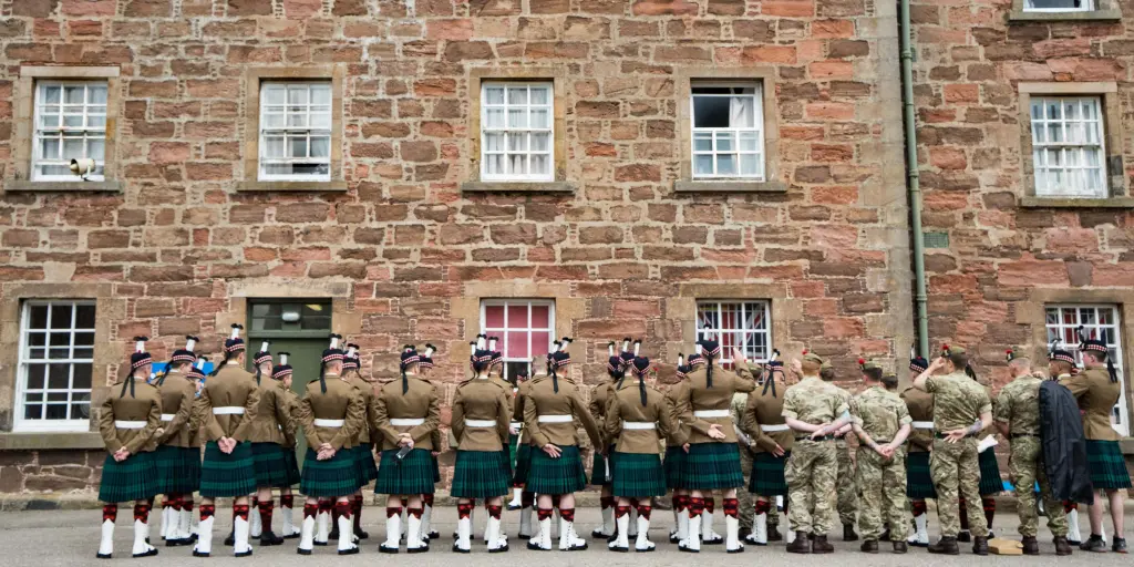 Soldiers wearing kilts and army fatigues with their backs to the camera outside a stone barracks building at Fort George, Scotland