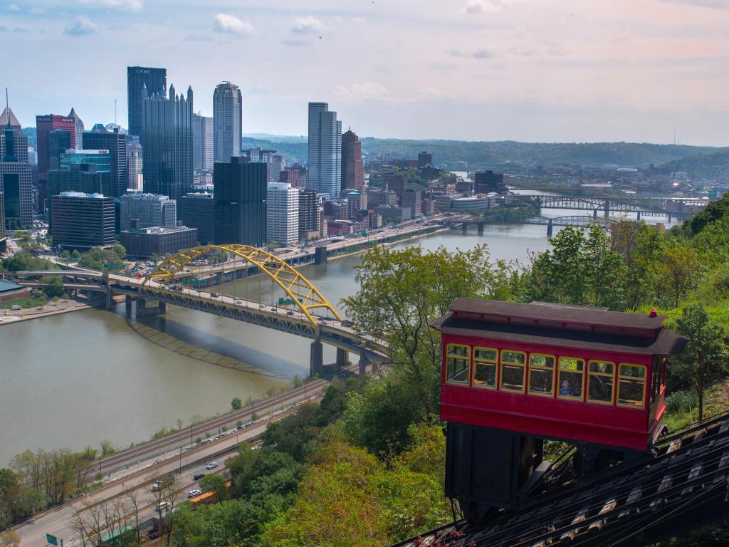 A red trolley car sits at the top of the sloping incline railroad track with a view over the river, bridges and downtown skyline