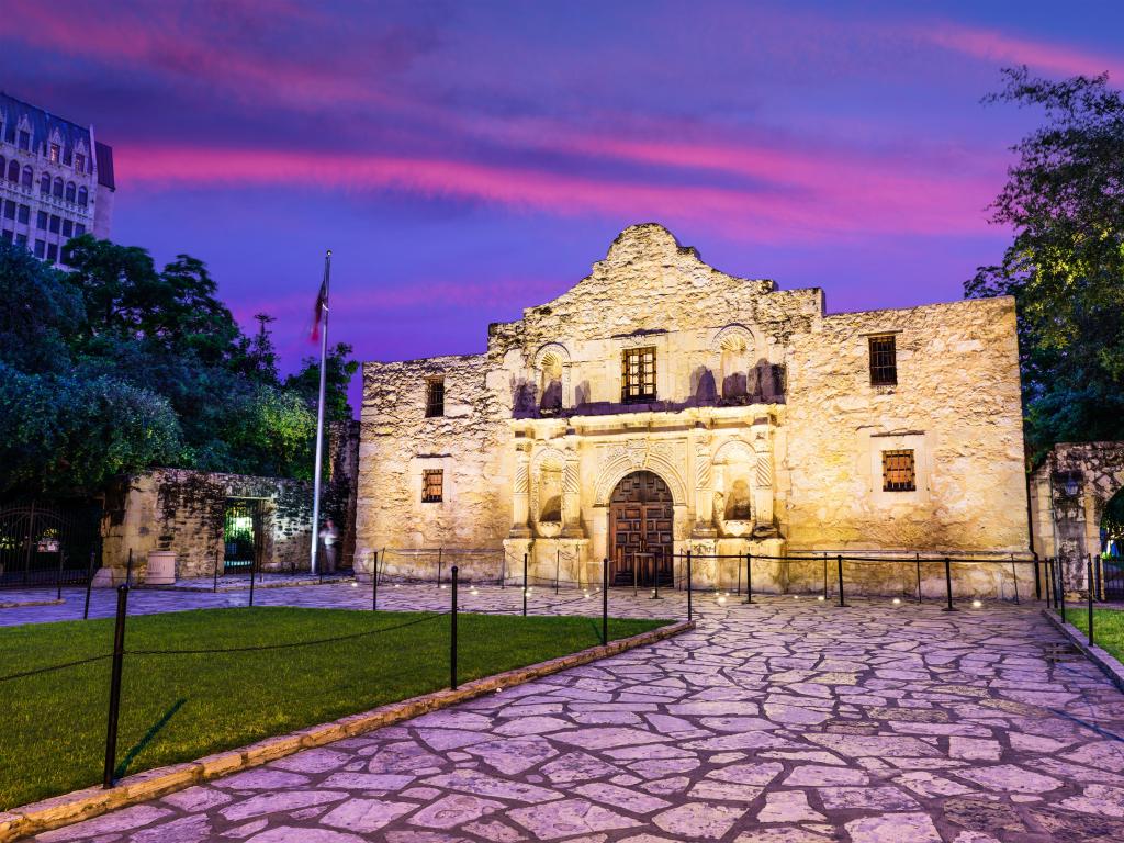 The Alamo is a famous landmark in San Antonio. The photo is taken during sunset with purple and pink clouds in the sky. A cobbled pathway leads to the lit-up landmark.