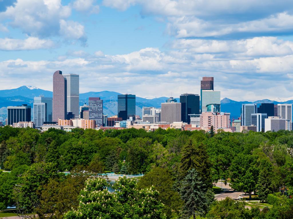 Denver, Colorado, USA with the city skyscrapers in the distance and mountains beyond, plus trees in the foreground on a sunny day.