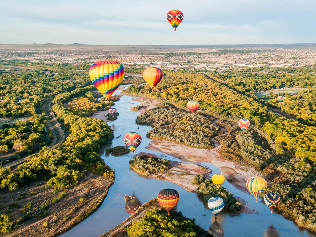 Albuquerque, New Mexico with hot air balloons flying over the Rio Grande river, with greenery below and the town in the distance. 
