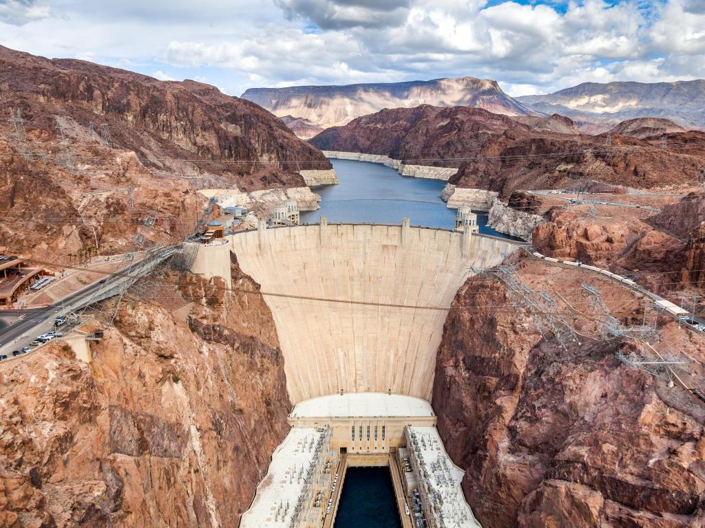 Hoover Dam, straddling Nevada and Arizona, USA taken as an aerial view with the red cliffs and Colorado River beyond, mountains in the distance under a cloudy and blue sky.