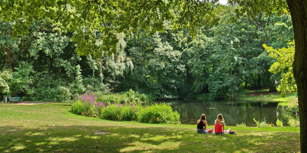 Two women sit on the grass and look out at a pond inside the lush green Tiergarten park in Berlin