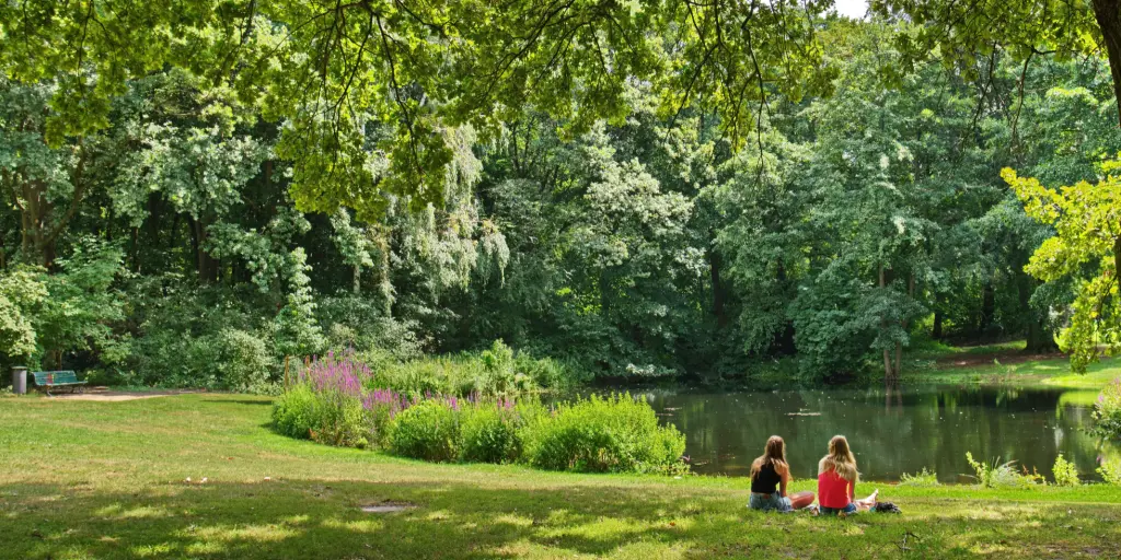 Two women sit on the grass and look out at a pond inside the lush green Tiergarten park in Berlin