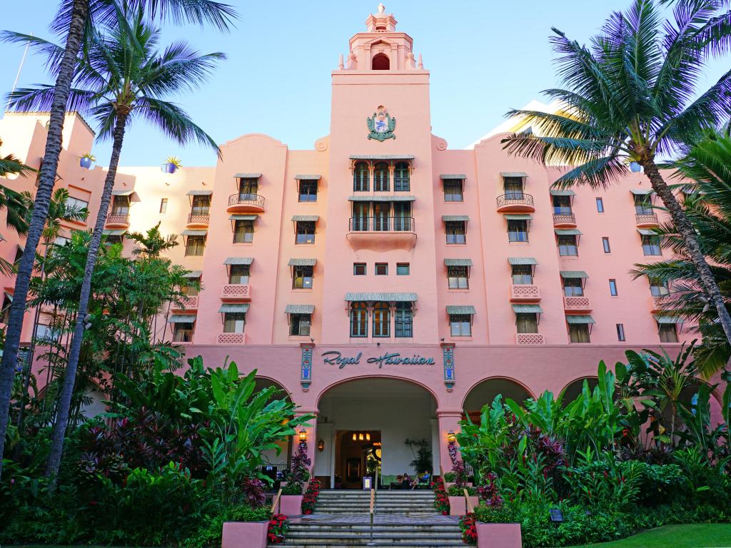 The front view of the famously pink hotel, the Royal Hawaiian, on a sunny day. Stairs leading to the entrance