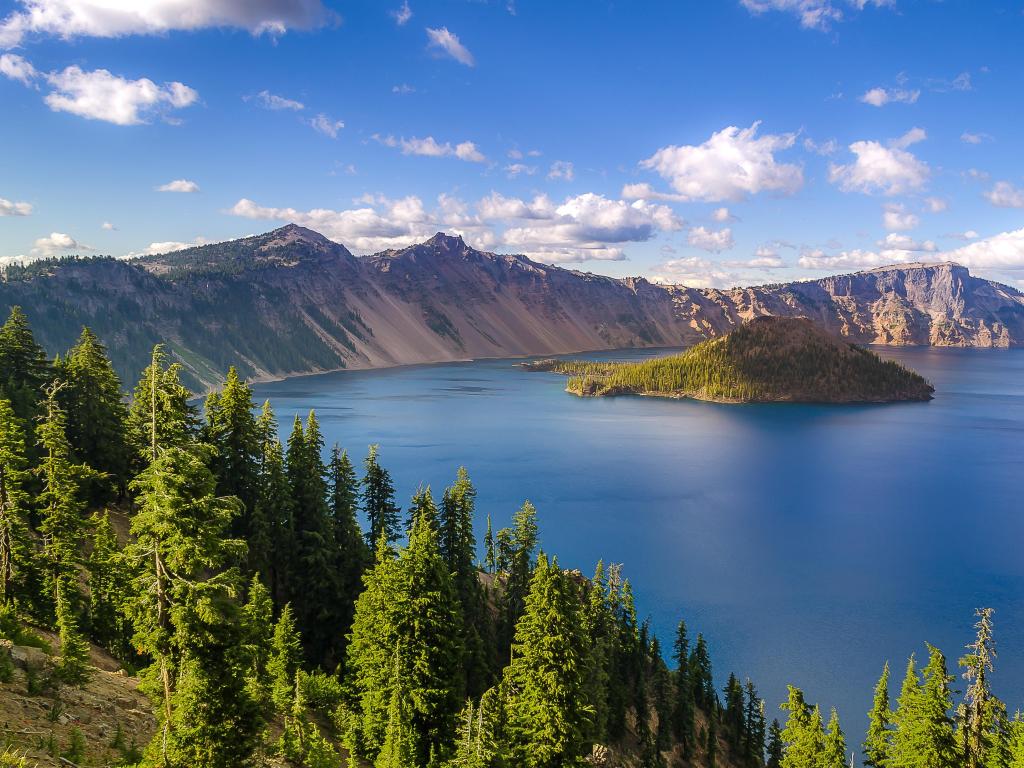 Crater Lake, Oregon, USA with a beautiful landscape of Crater Lake in summer with mountains in the distance and trees in the foreground.