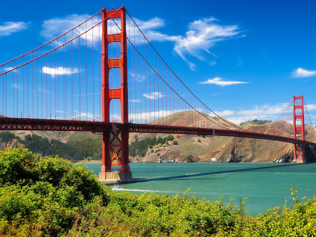 Red structure of the golden gate bridge crossing over turquoise water with blue sky and vivid green vegetation in the foreground