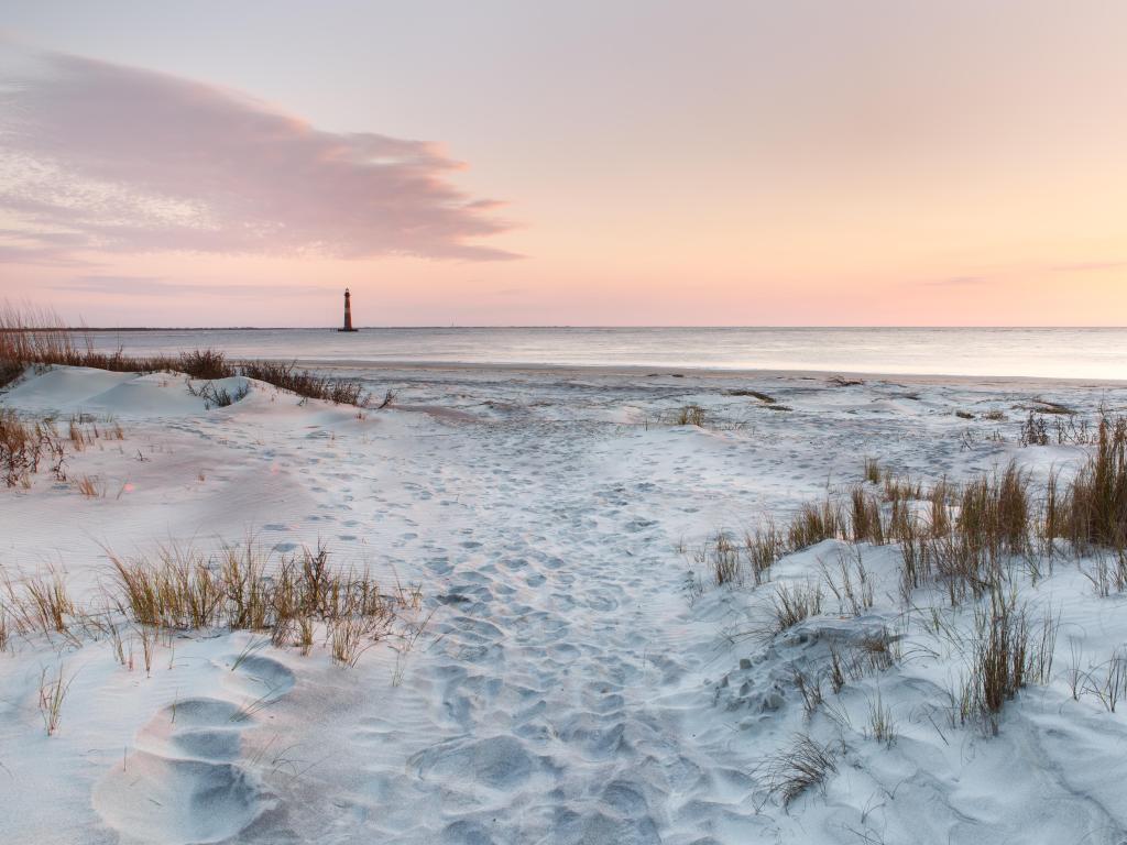 Folly Beach, Charleston, South Carolina, USA with sand and sea grasses on Folly Beach near Morris Island Lighthouse at sunset.