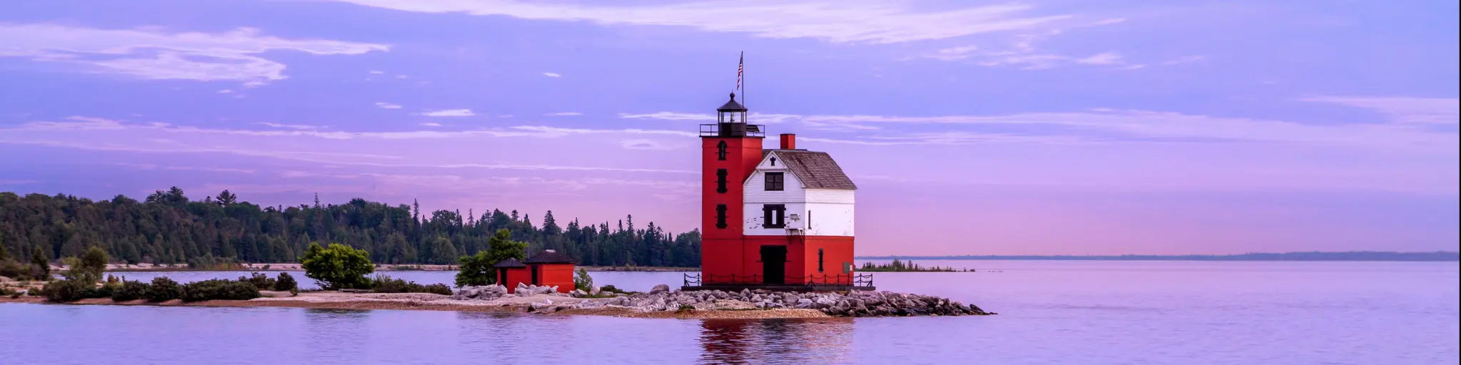 The historic Round Island Lighthouse at the shore of Mackinac Island in a purple and blue sky after sunset with a view of green pine trees at the distance